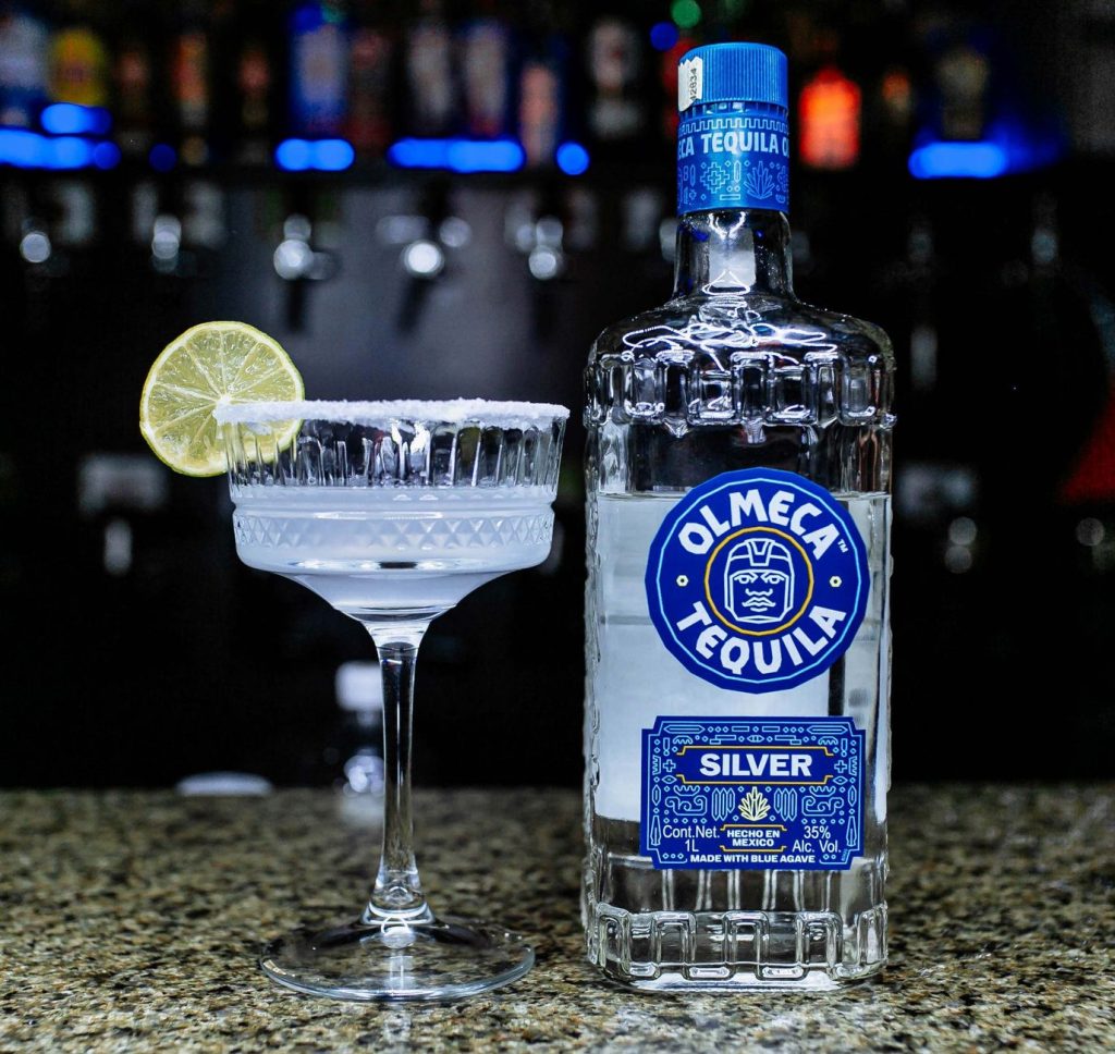 A margarita cocktail with a lime slice and Olmeca Tequila bottle on a bar counter.